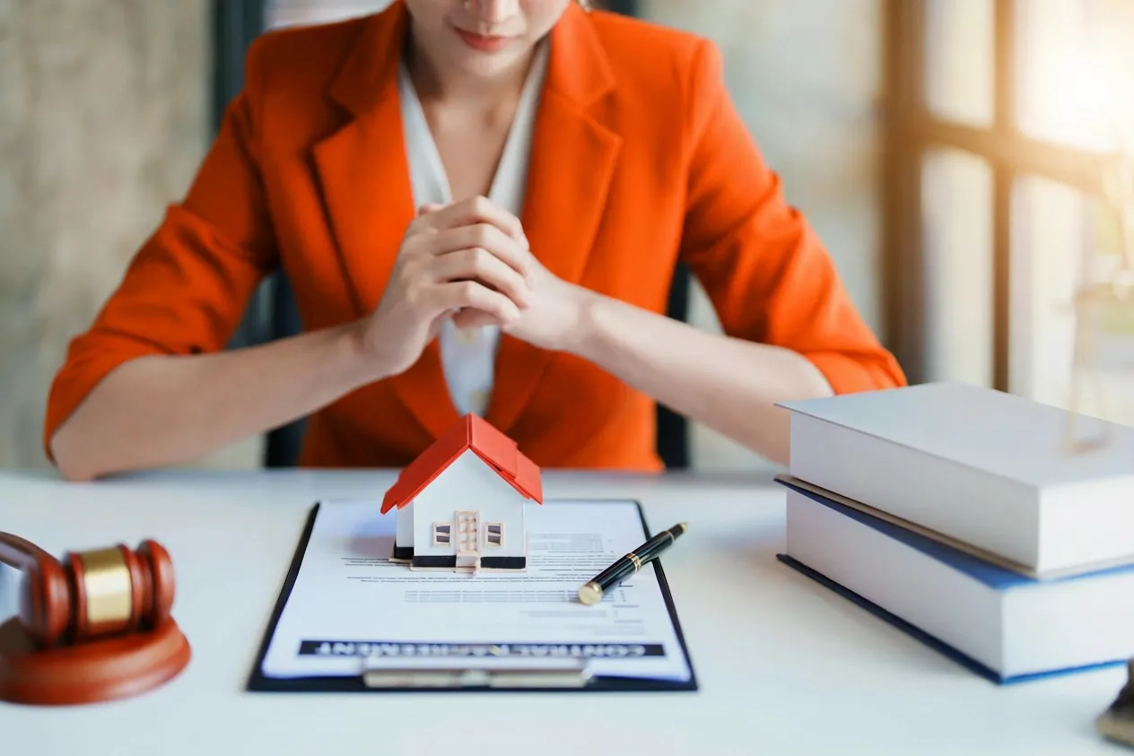 Professional in orange blazer with miniature house, contract, and gavel on desk.