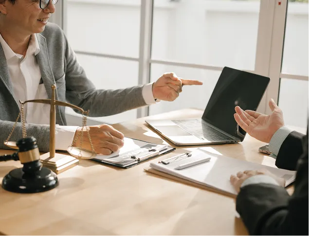 Two professionals in discussion at a desk with legal scales, a gavel, documents, and a laptop.