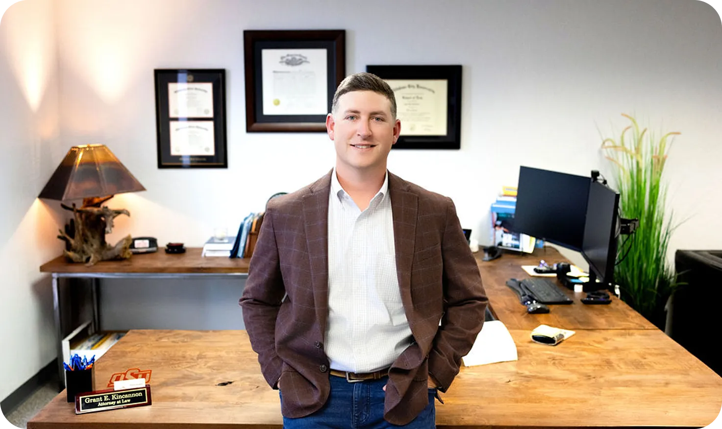 Professional man in blazer standing in office with wooden desk and framed diplomas.