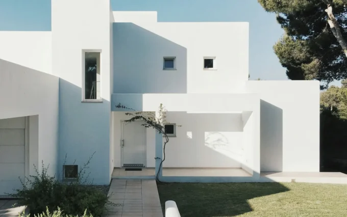 Modern white minimalist house exterior with geometric design, lawn, and surrounding greenery.