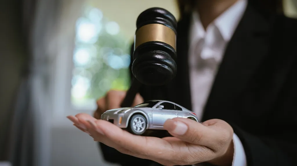 Person in formal attire holding a toy silver car and a judge's gavel above it.