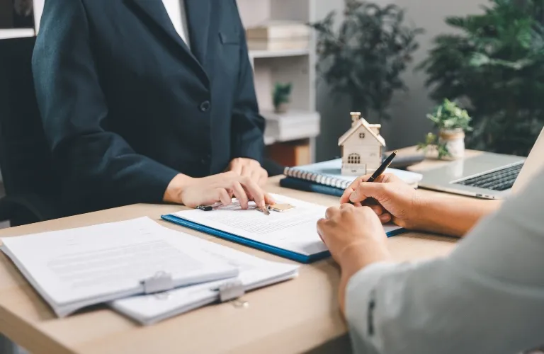 Two people engaged in signing documents at a desk with a miniature house model and laptop.