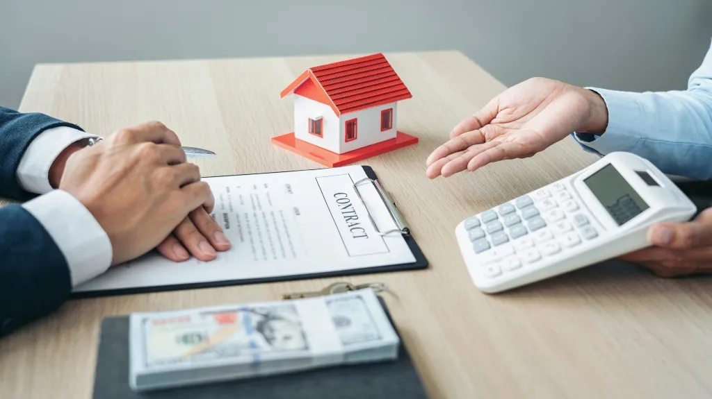 Two people discussing a contract with a model house, calculator, and money on a wooden table.