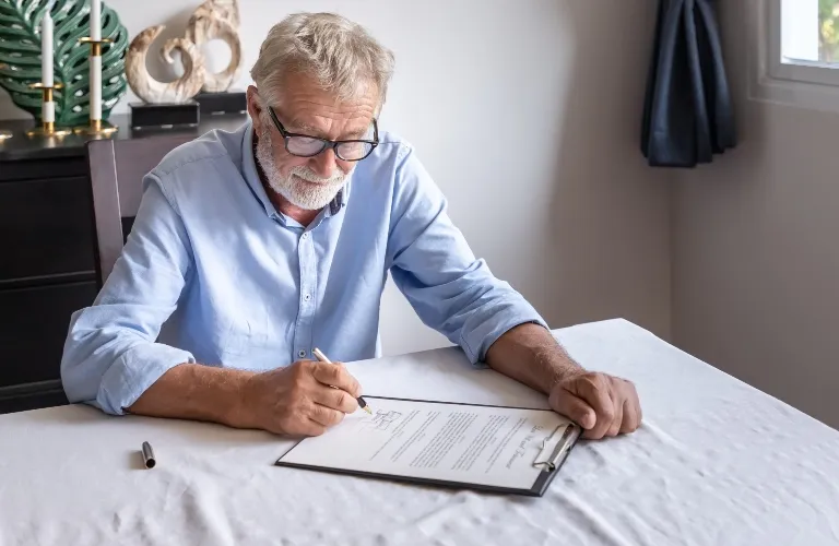Older man with glasses signing a document on a clipboard at a white table.
