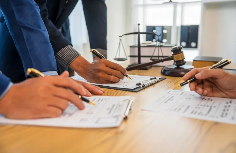 Three professionals in suits writing on documents at a wooden table with a judge’s gavel and scales of justice in the background.