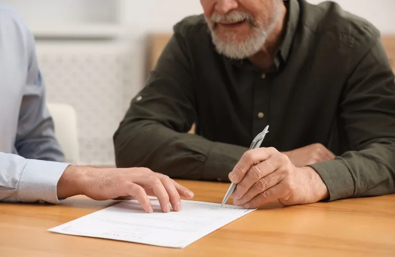 Two men at a wooden table reviewing and signing a document with a pen.