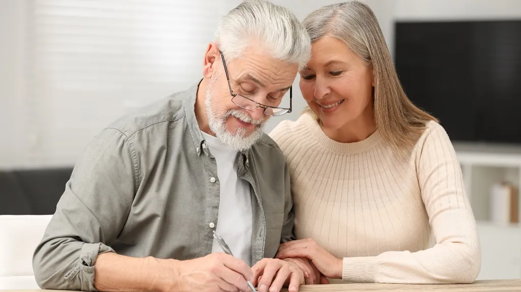 Mature man with gray hair signing document while woman in beige sweater supports him.