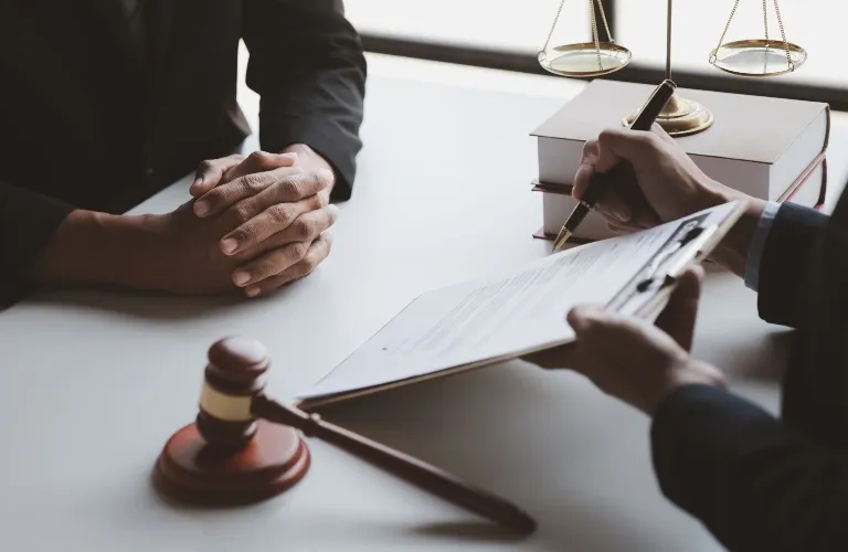Two individuals in a legal consultation, one signing documents, with gavel and scales on the desk.