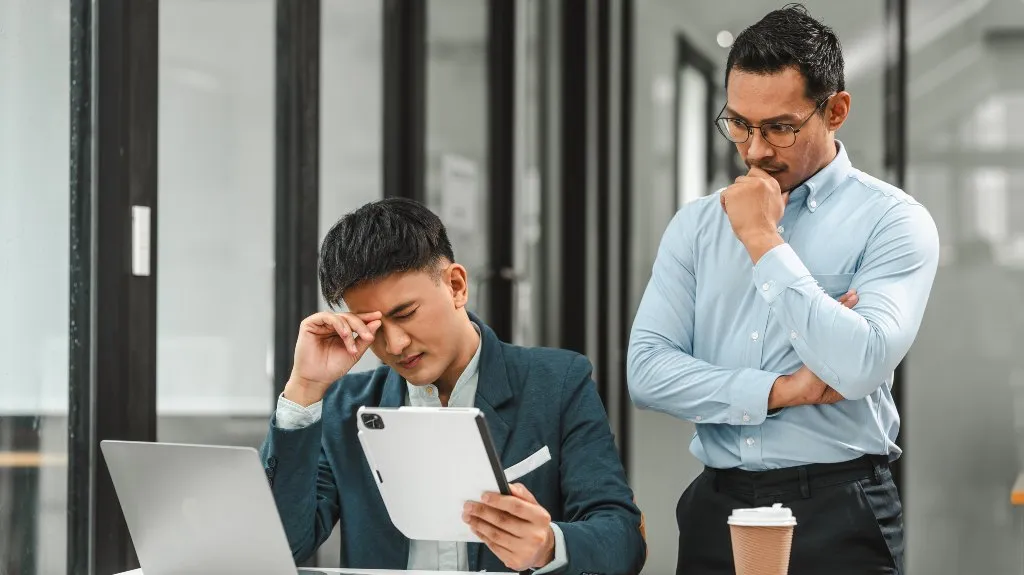Two businessmen in an office, one seated stressed holding a tablet, the other standing thoughtfully.