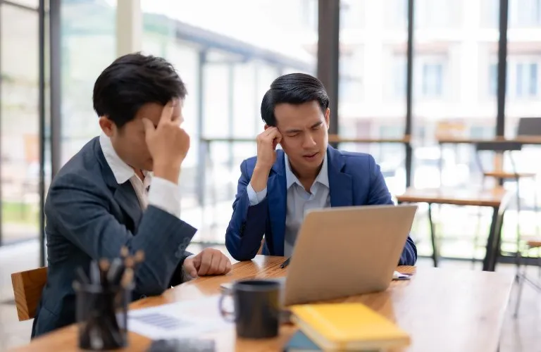 Two businessmen in suits appearing stressed while working on a laptop at a wooden table.