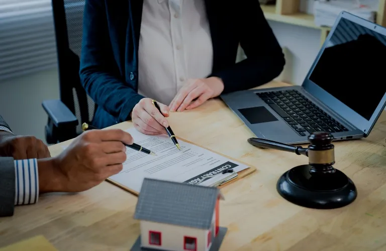 Two professionals reviewing a contract agreement at a desk with a laptop, gavel, and miniature house model.