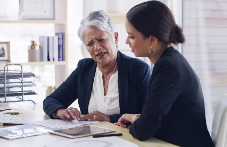 Two professional women in business attire engaged in a serious discussion at a desk with documents and a tablet.