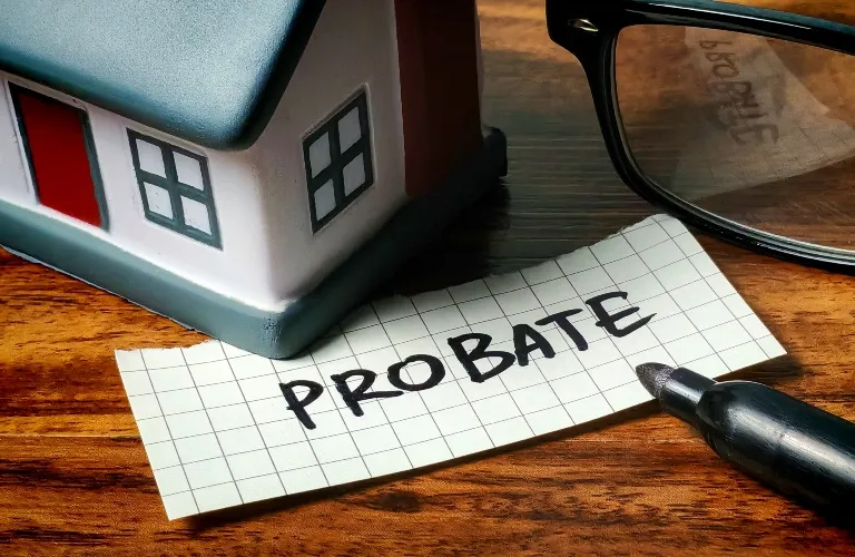 Small model house, handwritten "PROBATE" on graph paper, black marker, and eyeglasses on wooden surface.