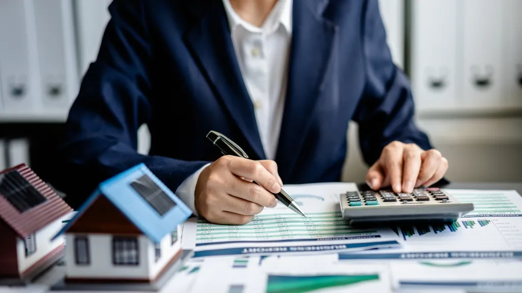 Person in a dark suit using a calculator and pen to analyze financial documents with model houses.