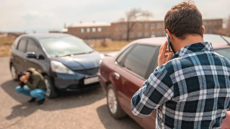 Man in plaid shirt talking on phone near two cars involved in a minor collision.