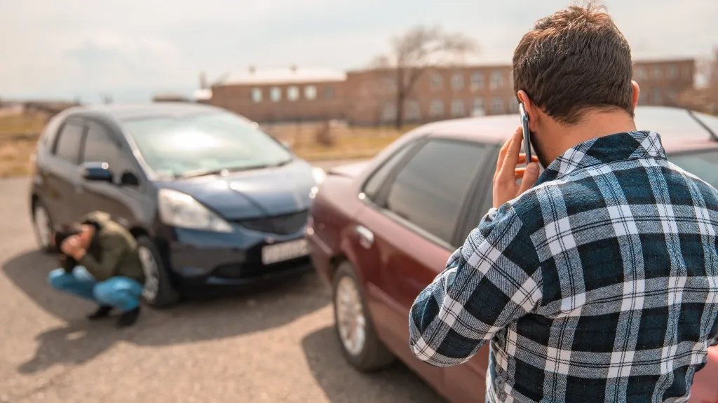Man in plaid shirt talking on phone near two cars involved in a minor collision.