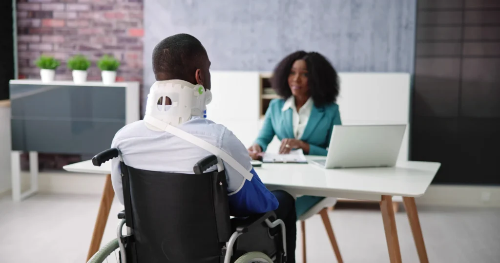 Man wearing a neck brace and arm sling in wheelchair consulting a professional woman at office desk.